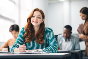 Modern Education Concept. Portrait of smiling ginger red haired female student sitting at desk in classroom at university, taking test or writing notes in her notebook, looking posing at camera