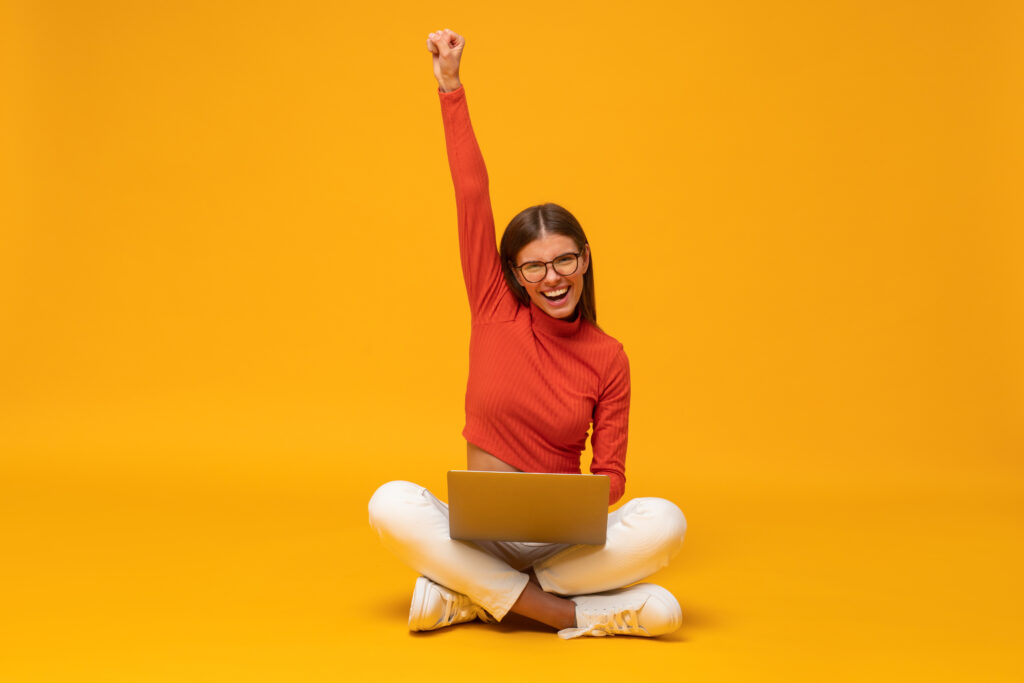 Winner. Excited happy business woman sitting on floor with laptop, raising one hand in the air saying yes, isolated on yellow background